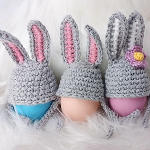 May include: Three gray crocheted bunny hats with pink and white ears. Each hat is decorated with a different colored egg inside.