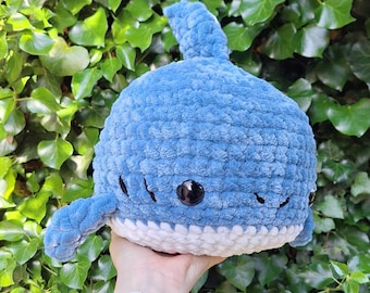 May include: A plush, blue crocheted shark with a white belly, black eyes, and small fins. The handmade toy is soft and cuddly, perfect for children or as a decorative item. The shark is set against a backdrop of green foliage.