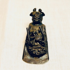 Puede incluir: Una figura plateada de una figura sonriente con cuernos, sosteniendo lo que parece ser un instrumento musical. La figura está sobre un fondo blanco. La figura tiene detalles intrincados y una apariencia desgastada.