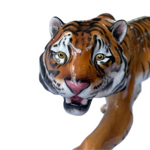 Op de afbeelding: Een keramieken tijgerbeeldje met een glanzende afwerking. De tijger heeft oranje en zwarte strepen, gele ogen en een roze mond. Het beeldje staat in een loopstand, met de mond iets open.