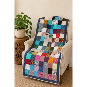 Könnte beinhalten: Eine Patchwork-Decke bestehend aus einem Raster aus bunten Stoffquadraten mit verschiedenen Mustern, darunter florale, geometrische und einfarbige Drucke. Die Decke ist mit einer dunkelblauen Stoffkante eingefasst und über einen hellen Sessel gelegt.