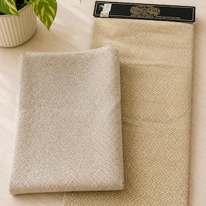 Op de afbeelding: Twee stukken beige stof met een subtiel visgraatpatroon. Het ene stuk is gevouwen, terwijl het andere plat ligt met een zwart label aan de bovenkant met de tekst Welsh Wool en Pure New Wool, en een klein zwart label in de onderste hoek.