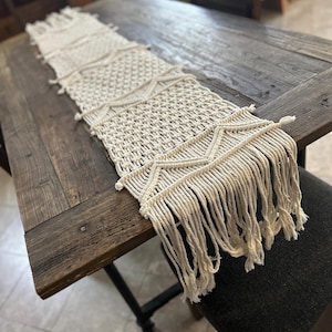 May include: Cream macrame table runner with geometric patterns and a fringed edge. The runner is displayed on a rustic wooden table. A decorative accent for dining or living spaces. The table runner measures approximately 150 cm in length.