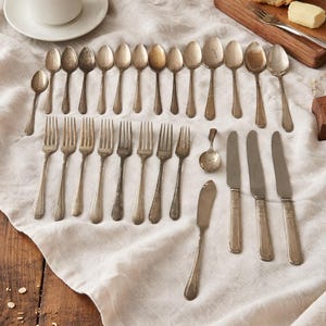 May include: A collection of antique silverware, including spoons, forks, and knives, laid out on a white linen cloth. A cup of coffee and a wooden board with bread and butter are also present, creating a rustic, elegant dining scene.