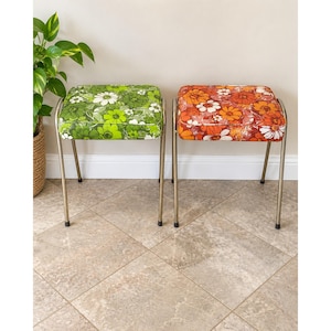May include: Two square stools with metal legs and padded seats. One seat features a green floral pattern, while the other features an orange and white floral pattern. Both stools have white piping along the edges of the seat cushions.