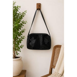 Peut inclure: Un sac à bandoulière noir présentant un motif en patchwork composé de panneaux de cuir lisse et texturé. Ce sac rectangulaire est doté d'une longue bandoulière unique et est suspendu à un crochet mural en bois. La surface présente des formes géométriques variées.