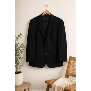 Könnte beinhalten: Ein einfarbiger schwarzer Blazer mit steigendem Revers, einer Knopfleiste mit drei Knöpfen und zwei Pattentaschen an der Taille. Das Kleidungsstück hängt an einem Holzkleiderbügel und hat lange Ärmel mit jeweils vier kleinen Knöpfen.
