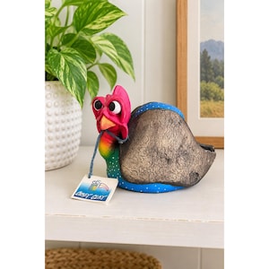 May include: A turkey figurine with a bright pink head, orange beak, and large white eyes. The body features a textured bronze-colored back, a blue chest with white polka dots, and a rainbow-colored neck. A tag attached to the beak reads Crazy Clay.