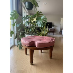 May include: A mid-century modern style ottoman with a pink, clover-shaped cushion. The cushion is button-tufted and sits atop a round, wooden base with tapered legs. The legs have brass-colored tips. The ottoman is a decorative furniture piece.