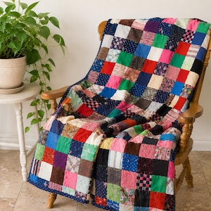 Peut inclure: Une courtepointe en patchwork composée d'une grille de carrés de tissu multicolores aux motifs et couleurs variés, incluant le rouge, le bleu, le vert, le rose, le blanc et le noir, drapée sur une chaise à bascule en bois à haut dossier.