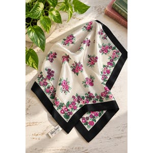 May include: A square silk scarf featuring a white background with a repeating pattern of small pink flowers and green leaves. The scarf is framed by a solid black border along the edges and includes a small white fabric tag with black text.