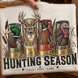 May include: A cream-coloured sweatshirt featuring a graphic of four shotgun shells, each containing a different animal: a mallard duck, a deer, a pheasant, and a turkey. Below the shells, the text reads HUNTING SEASON FIELD FOWL GAME.