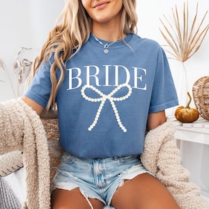 Może przedstawiać: Niebieska koszulka z napisem "BRIDE" w białym skrypcie i perłową kokardą. Koszulka jest z krótkim rękawem i ma luźny krój. Modelka ma na sobie dżinsowe szorty i kremowy kardigan.