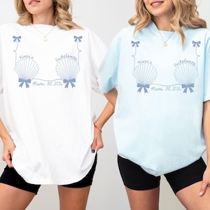 Puede incluir: Camisetas blancas y celeste con un diseño de conchas marinas y el texto "Kiara's Bachelorette Miami, FL 2026". Las camisetas tienen mangas cortas y un corte holgado, ideales para una despedida de soltera.