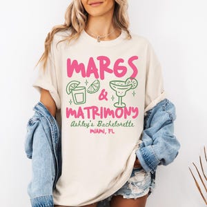 May include: A cream-colored t-shirt featuring the pink text MARGS & MATRIMONY, green line art of a cocktail glass and lime wedge, and the text Ashley's Bachelorette MIAMI, FL in green and pink, layered under a light blue denim jacket.
