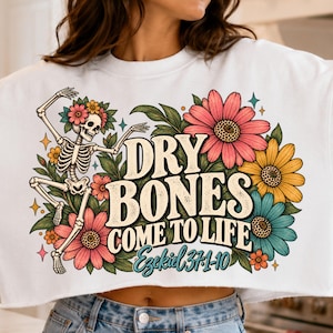 Könnte beinhalten: Ein weißes bauchfreies Sweatshirt mit einer Grafik eines tanzenden Skeletts, umgeben von rosa, gelben und türkisfarbenen Blumen. Der Text "DRY BONES COME TO LIFE" ist in fetter Schrift gedruckt, darunter steht "Ezekiel 37:1-10".