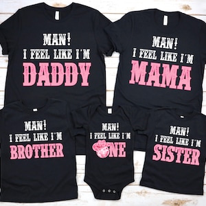 以下が含まれることがあります： 白とピンクの文字が入った黒いTシャツ。「MAN！I FEEL LIKE I'M DADDY」、「MAN！I FEEL LIKE I'M MAMA」、「MAN！I FEEL LIKE I'M BROTHER」、「MAN！I FEEL LIKE I'M ONE」、そして「MAN！I FEEL LIKE I'M SISTER」と書かれています。