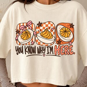 Puede incluir: Una camiseta color crema con un gráfico de tres huevos rellenos dentro de cuadrados a cuadros naranjas y blancos, decorada con lazos rosas y estrellas. El texto "YOU KNOW WHY I'M HERE." está impreso en letras negras y a cuadros naranjas.