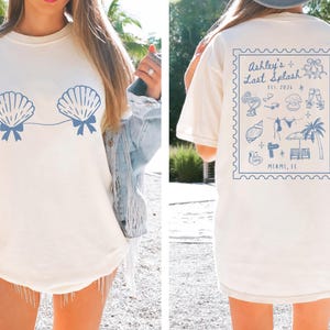 May include: Cream-colored t-shirt with a blue seashell design on the front and a blue stamp-like graphic on the back. The front features two seashell shapes connected by a line. The back graphic says "Ashley's Last Splash" and "Miami, FL".