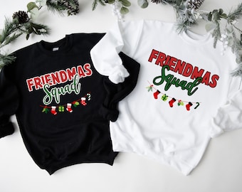 May include: Two sweatshirts, one black and one white, with the text "FRIENDMAS Squad" in red and green. A string of Christmas stockings and gifts is below the text. The sweatshirts are likely for a holiday or Christmas theme.