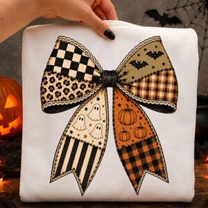 May include: A white sweatshirt featuring a large, central graphic of a decorative bow. The bow is composed of various patterned segments including black and white checkers, leopard print, bats, ghosts, pumpkins, and orange and black plaid.