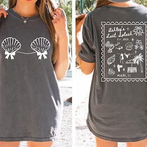 May include: Dark gray t-shirt featuring a white seashell design on the front. The back of the shirt has a white graphic with the text "Ashley's Last Splash" and various beach-themed illustrations. The text "Miami, FL" is also included.