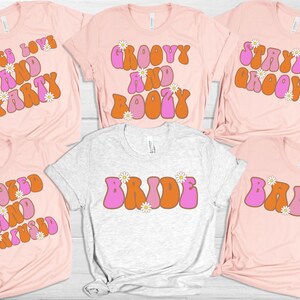Könnte beinhalten: Sechs rosafarbene und weiße T-Shirts mit groovigem Text und Blumendesigns. Die Shirts sagen "Peace Love and Party", "Groovy and Boozy", "Stayin' Groovy", "Booked and CooCoo", "Bride" und "Babe".