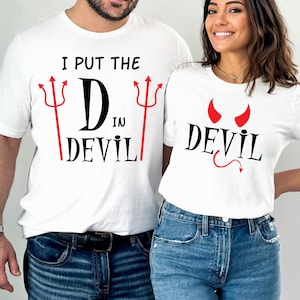 May include: White t-shirts with black and red devil-themed graphics. One shirt reads "I PUT THE D IN DEVIL" with red trident accents. The other shirt says "DEVIL" with red horns and a tail.
