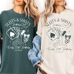 May include: Two t-shirts, one dark green and one cream, featuring a graphic design with the text "SLOTS & SHOTS" and "Birthday Girl" or "Birthday Crew". The design includes playing cards, a martini, dice, and cherries. The text "Emily's 21st Birthday" and "Las Vegas" are also present.