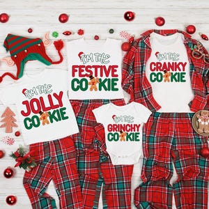 May include: A collection of Christmas pajamas with matching tops and bottoms. The white tops have the phrases "I'm the Jolly Cookie", "I'm the Festive Cookie", "I'm the Grinchy Cookie", and "I'm the Cranky Cookie" printed on them. The bottoms are red and green plaid.