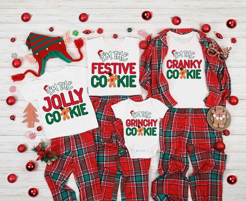 May include: A collection of Christmas pajamas with matching tops and bottoms. The white tops have the phrases "I'm the Jolly Cookie", "I'm the Festive Cookie", "I'm the Grinchy Cookie", and "I'm the Cranky Cookie" printed on them. The bottoms are red and green plaid.