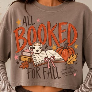 May include: A grey short-sleeved t-shirt featuring a graphic with the text ALL BOOKED FOR FALL and just one more chapter. The design includes an open book with a pink ribbon, a coffee mug, a pumpkin, autumn leaves, and small stars.