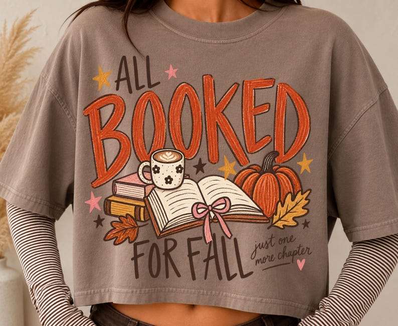 May include: A grey short-sleeved t-shirt featuring a graphic with the text ALL BOOKED FOR FALL and just one more chapter. The design includes an open book with a pink ribbon, a coffee mug, a pumpkin, autumn leaves, and small stars.