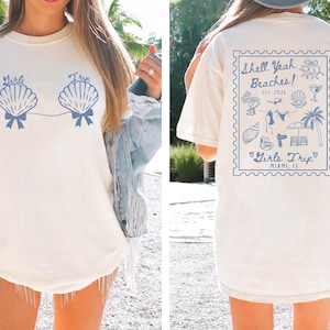 May include: Cream-colored t-shirt with blue seashell graphic and text "Girls Trip". The back of the shirt features a blue stamp design with the text "Shell Yeah Beaches! Est. 2024" and "Girls Trip Miami, FL".