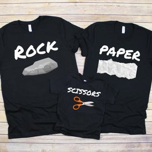 Puede incluir: Camisetas negras con texto y gráficos blancos. Una camiseta dice "ROCK" con un gráfico de roca, otra dice "PAPER" con un gráfico de papel y la tercera dice "SCISSORS" con un gráfico de tijeras.