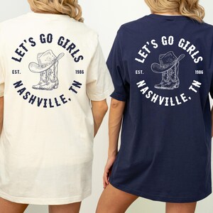 May include: Two t-shirts, one cream and one navy blue, feature a circular graphic on the back. The graphic includes a cowboy hat and boots, with the text "LET'S GO GIRLS NASHVILLE, TN" and the year "1986".