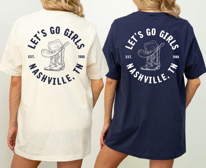 May include: Two t-shirts, one cream and one navy blue, feature a circular graphic on the back. The graphic includes a cowboy hat and boots, with the text "LET'S GO GIRLS NASHVILLE, TN" and the year "1986".