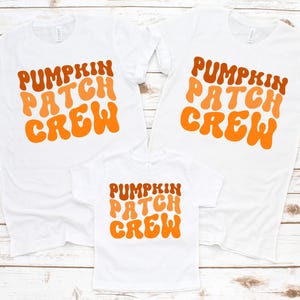 May include: Three white t-shirts with the text "PUMPKIN PATCH CREW" in orange and brown retro font. The shirts are short-sleeved and appear to be made of cotton. The shirts are arranged in a family-like grouping.