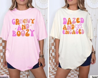 May include: Two pink and white oversized t-shirts with a retro graphic design. The pink shirt says "Groovy and Boozy" and the white shirt says "Dazed and Engaged".