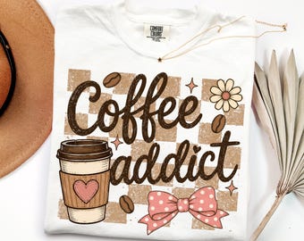 May include: A white t-shirt featuring a brown checkered background with the text Coffee addict in dark brown script. The design includes a coffee cup with a pink heart, a pink polka dot bow, coffee beans, a flower, and small star accents.