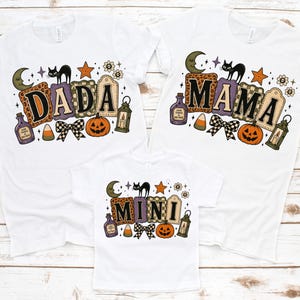 Può includere: Tre magliette bianche con grafiche a tema Halloween. Ogni maglietta mostra le parole DADA, MAMA o MINI in stampatello, circondate da illustrazioni di un gatto nero, luna crescente, zucca, lanterna, pozione e un fiocco.