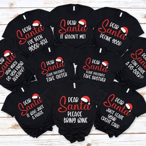 Puede incluir: Una colección de camisetas negras con varias frases navideñas humorísticas en rojo y blanco. Cada camiseta presenta las palabras "Dear Santa" y un gráfico de gorro de Papá Noel, seguido de un mensaje único.