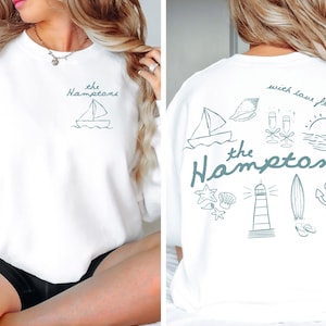 May include: White sweatshirt with teal text and illustrations. The front features "the Hamptons" and a sailboat. The back displays "with love from, the Hamptons" with nautical images like a lighthouse, anchor, and surfboard.