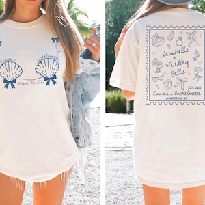 May include: A cream-coloured t-shirt with a blue seashell design and text reading "Lauren's Bachelorette Miami, FL 2026". The back features a stamp-like design with "Seashells + Wedding Bells" and "Lauren's Bachelorette Charleston, SC".