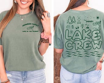May include: Sage green t-shirt with "JOHNSON FAMILY LAKE OF THE OZARKS" printed on the front. The back of the shirt features the text "LAKE CREW" with lake-themed illustrations, including trees, a campfire, and a kayak.