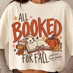 May include: An off-white short-sleeve t-shirt featuring a graphic with the text ALL BOOKED FOR FALL and just one more chapter. The design includes an open book, a coffee mug, a pumpkin, autumn leaves, and stars in orange, pink, and brown.