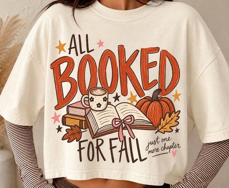 May include: An off-white short-sleeve t-shirt featuring a graphic with the text ALL BOOKED FOR FALL and just one more chapter. The design includes an open book, a coffee mug, a pumpkin, autumn leaves, and stars in orange, pink, and brown.