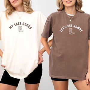May include: Two t-shirts, one cream and one brown, with rodeo-themed text and a boot graphic. The cream shirt says "MY LAST RODEO" and the brown shirt says "LET'S GET ROWDY". Both shirts are short-sleeved and have a relaxed fit.