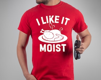 Pode incluir: Camiseta vermelha com o texto branco "I LIKE IT MOIST" e um gráfico de um peru assado em um prato. A camiseta é de manga curta.