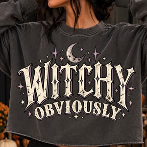 May include: A charcoal gray long-sleeve cropped sweatshirt featuring a distressed white graphic print that reads "WITCHY OBVIOUSLY" in a stylized font, accented by a crescent moon and small purple stars surrounding the text.